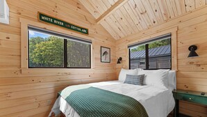 A wooden cabin bedroom with a bed, two windows, a wall-mounted lamp, and a sign indicating the White River COTTER. ARK.