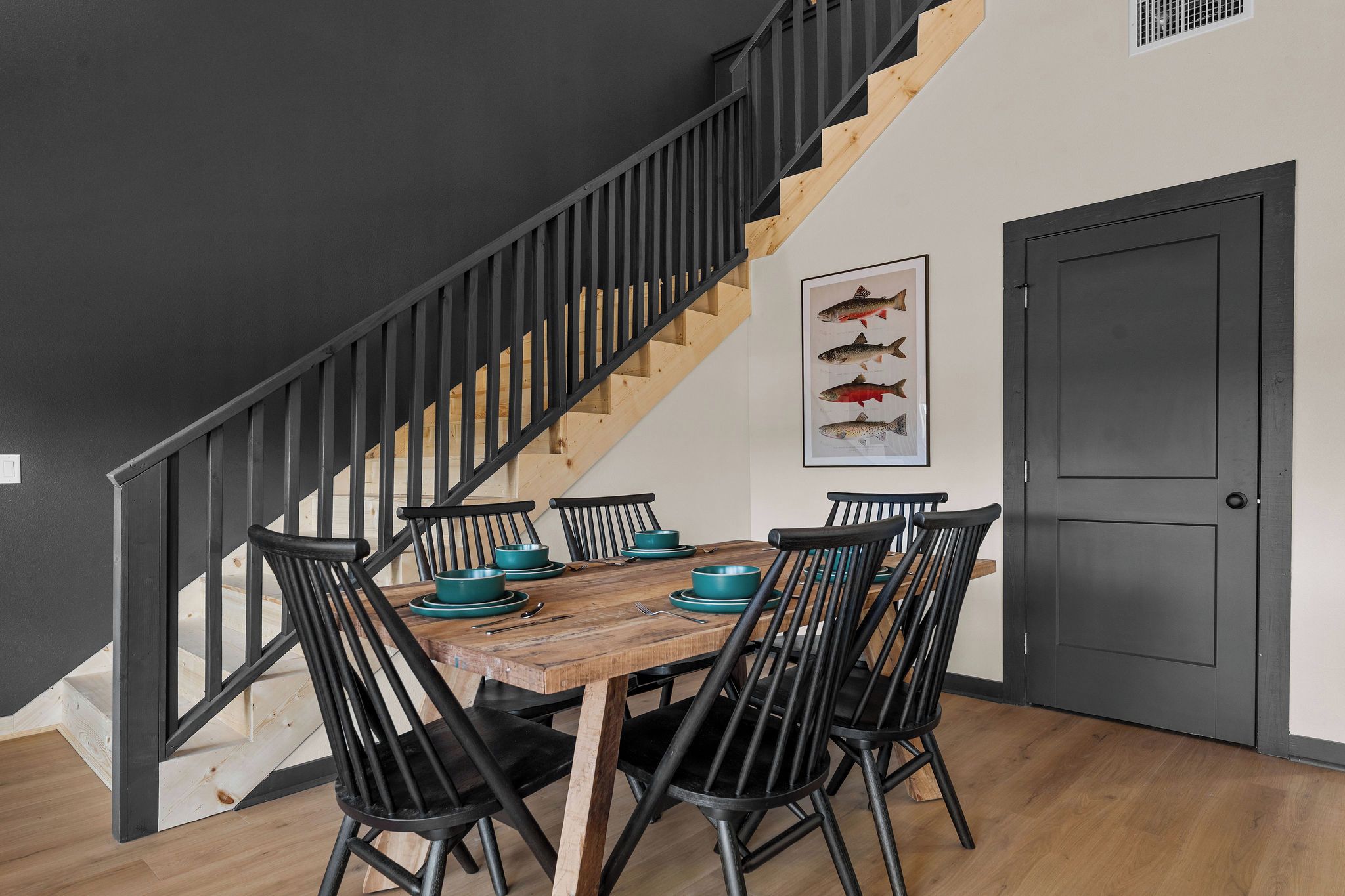 Een eetgedeelte met een houten tafel, zwarte stoelen en teal kommen. Een trap met zwarte leuningen leidt naar een overloop.