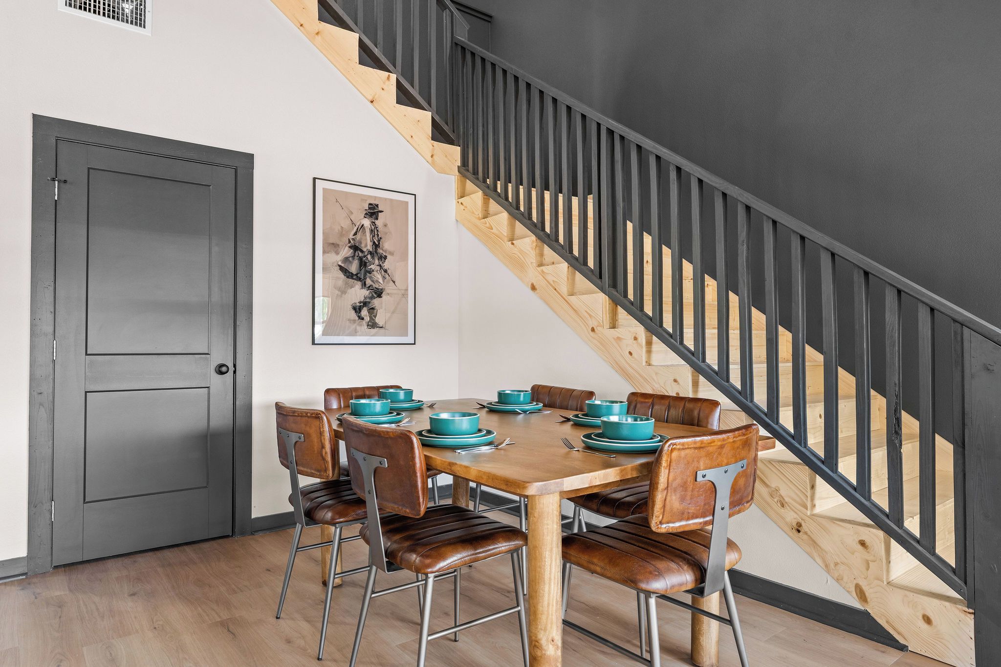 A dining area with a wooden table set for six, surrounded by leather chairs, a staircase with wooden steps and black railings, and a grey door.