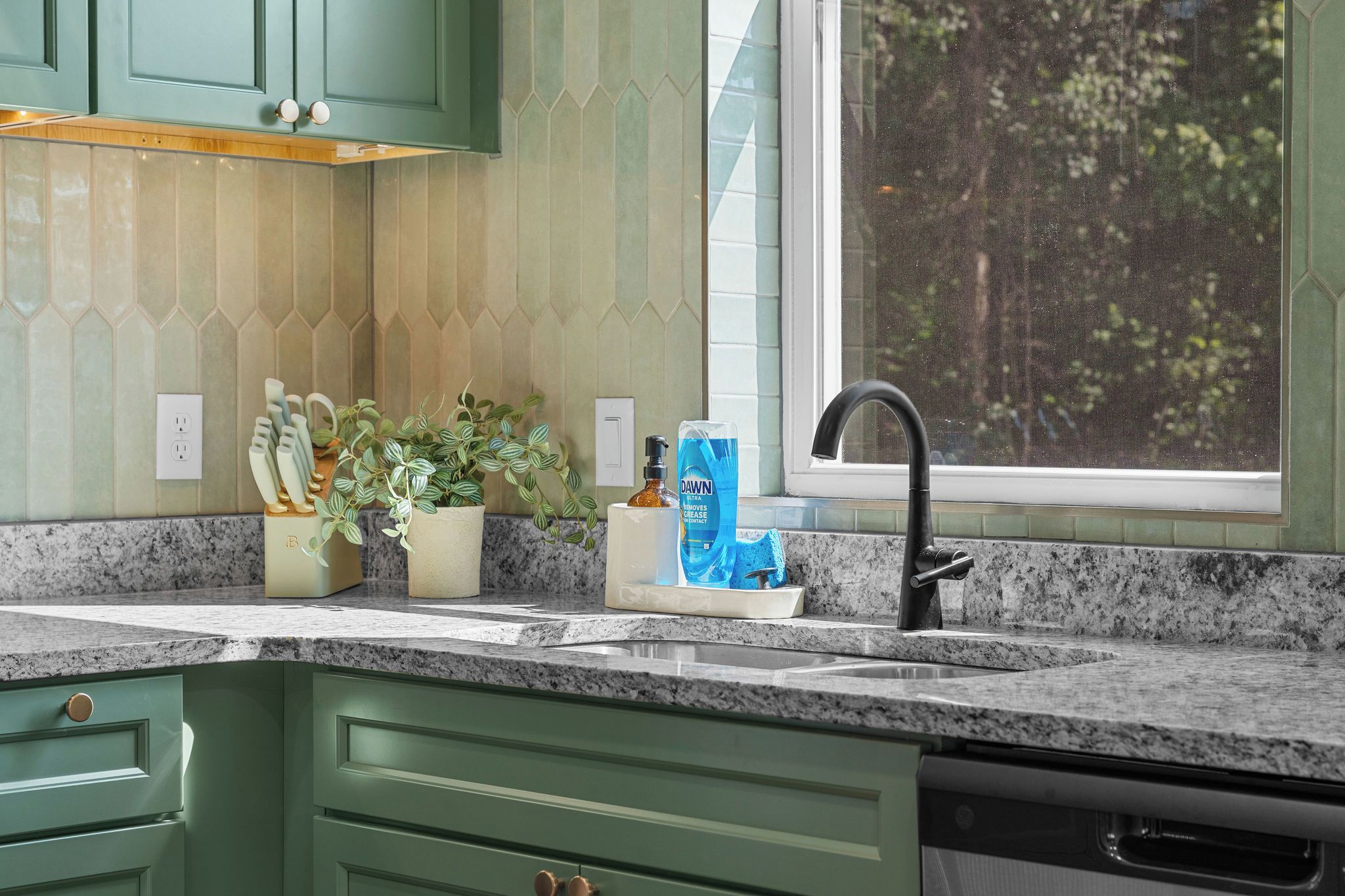 A kitchen with green cabinets, a granite countertop, a window with a view of trees, and a faucet.