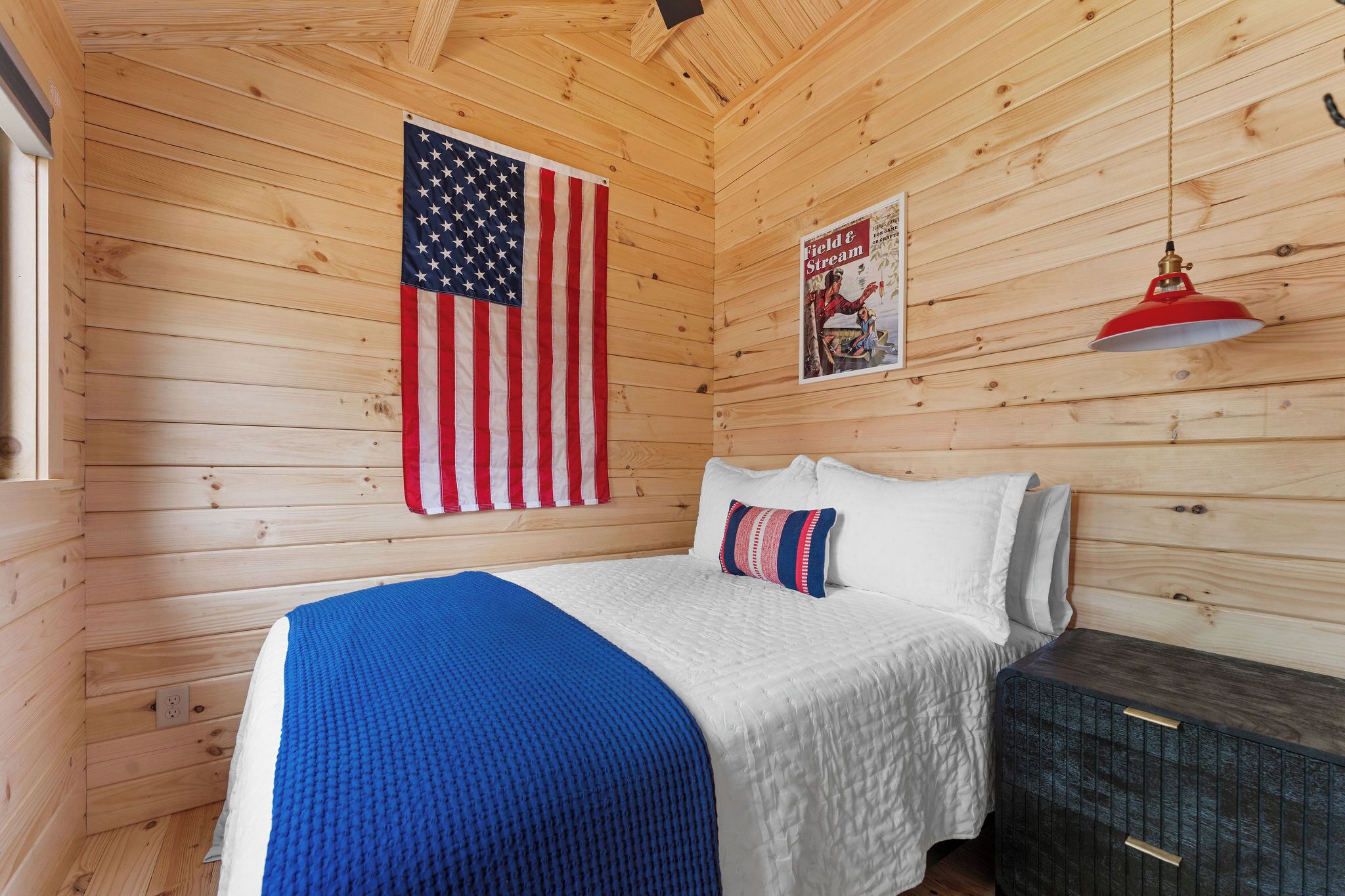 A wooden cabin room with a bed, a flag, a poster, and a red lamp.