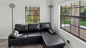 A modern living room with a black leather sectional sofa, two tall floor lamps, and large windows with a view of greenery. - Brand New Private Guest House (Miami)