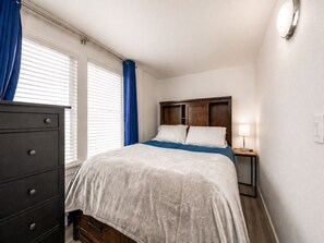 Un dormitorio con cama, cómoda, mesita de noche, lámpara y ventana con cortinas.