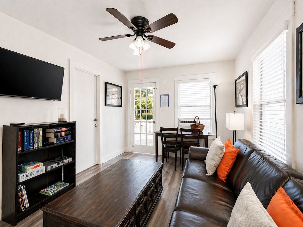 A living room with a dark wooden coffee table, a leather sofa with orange and white cushions, a bookshelf, a flat-screen TV, a ceiling fan, and a dining area with a table and chairs.