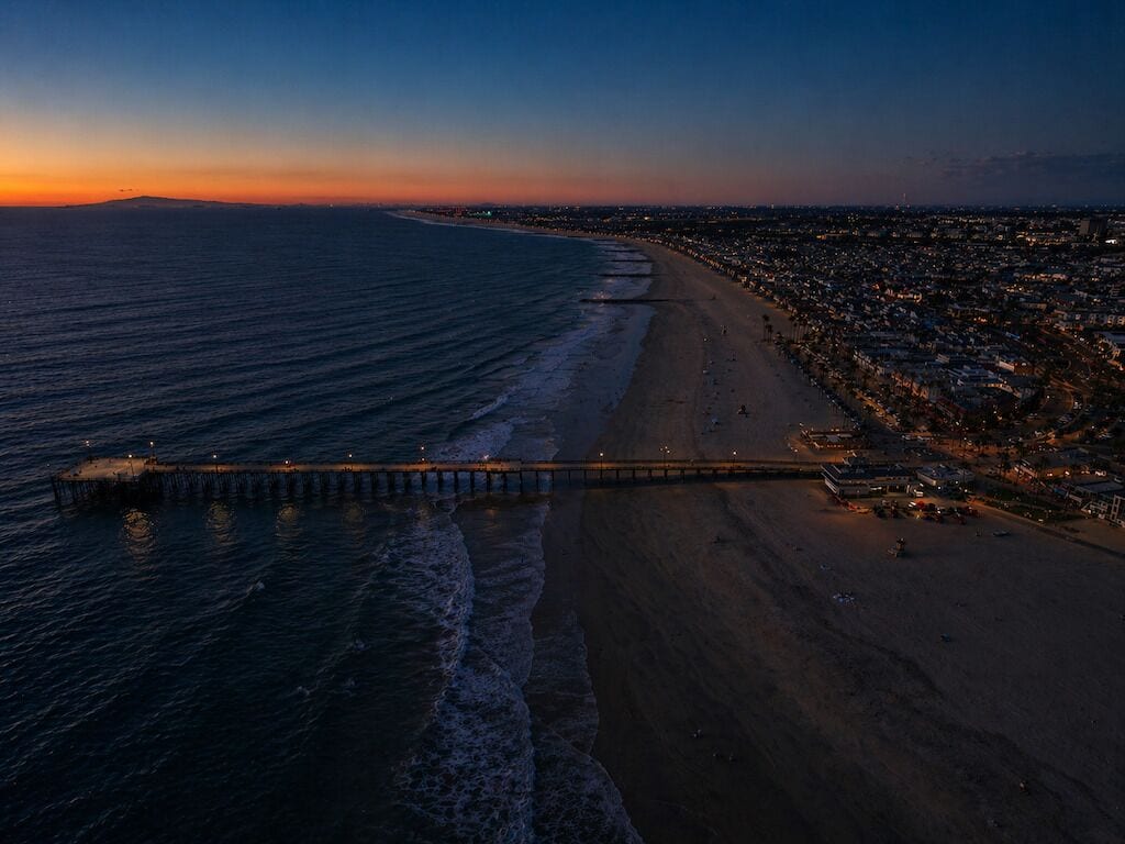 Une vue aérienne d’une plage avec une jetée qui s’avance dans l’eau, un paysage urbain en arrière-plan et un ciel au coucher du soleil.