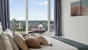 Ein Schlafzimmer mit großem Fenster, einem Bett mit Kissen und Blick auf eine Stadtlandschaft.