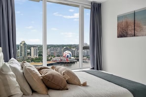 Un dormitorio con un ventanal grande, una cama con almohadas y vistas a un paisaje urbano.