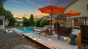 Un área junto a la piscina con una terraza de madera, sillas de descanso, una jaula decorativa y luces colgantes.