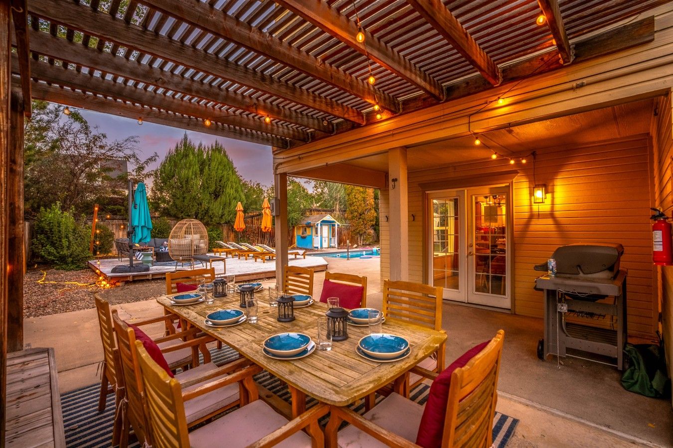 Un patio al aire libre con mesa de madera dispuesta para comer, rodeado de sillas, y una zona de piscina al fondo.