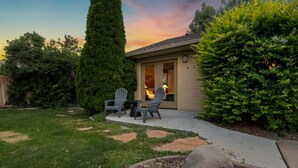 Una propiedad residencial con un patio, dos sillas Adirondack, un árbol conífero alto y un césped bien cuidado.