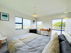 Ein Schlafzimmer mit einem großen Bett, einem Nachttisch, einem Sessel und zwei Fenstern mit Blick ins Freie.