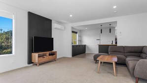 Een moderne woonkamer met een flatscreen televisie, een grijze bank, een houten salontafel en een keuken in de achtergrond.