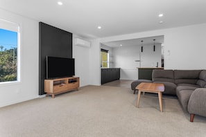 A modern living room with a flat-screen TV, a grey sofa, a wooden coffee table, and a kitchen area in the background. - Ocean Breeze Escape - Ohope Holiday Home (Ohope)