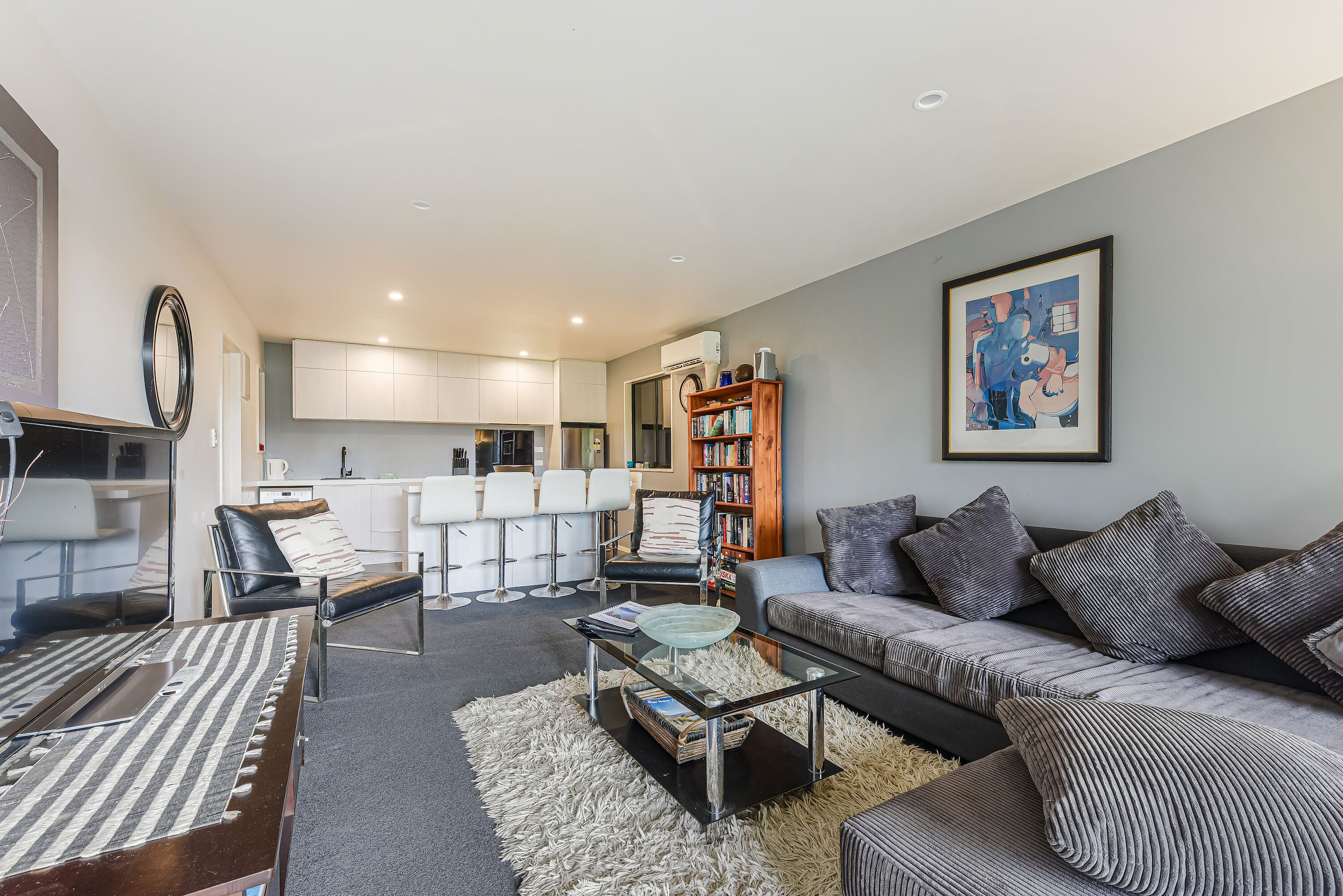 A modern living room with a sofa, coffee table, and chairs, a kitchen in the background, and a bookshelf.