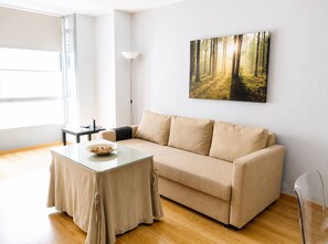 Ein modernes Wohnzimmer mit einem beige farbenen Sofa, einem Glastisch, einer Stehlampe und einer Wandkunst mit Waldmotiv.