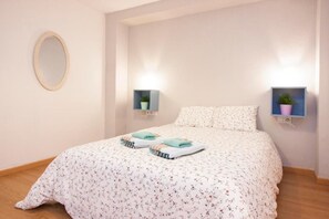 Ein Schlafzimmer mit floralem Bettbezug, zwei Nachttischen mit Topfpflanzen, einem runden Spiegel an der Wand und einem Holzboden.