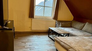 Una habitación con piso de madera, un sofá, un banco, un televisor y una ventana con cortinas.
