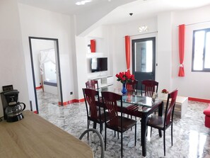 Un coin repas avec une table en verre, des chaises rouges, un téléviseur fixé au mur et un rideau rouge.