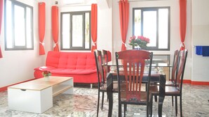 Een kamer met een rode bank, een eettafel met stoelen, een salontafel en een raam met gordijnen.