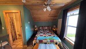 Um quarto aconchegante com teto de madeira, uma cama com colcha colorida, luminárias de cabeceira, uma janela com cortinas e um banco de madeira.