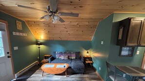 Um quarto aconchegante com um ventilador de teto, um sofá, uma mesinha, uma cadeira e um abajur.