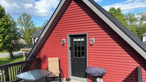 Um edifício vermelho com porta preta, um pequeno alpendre com mesa e cadeiras, e um carrinho de bebê.
