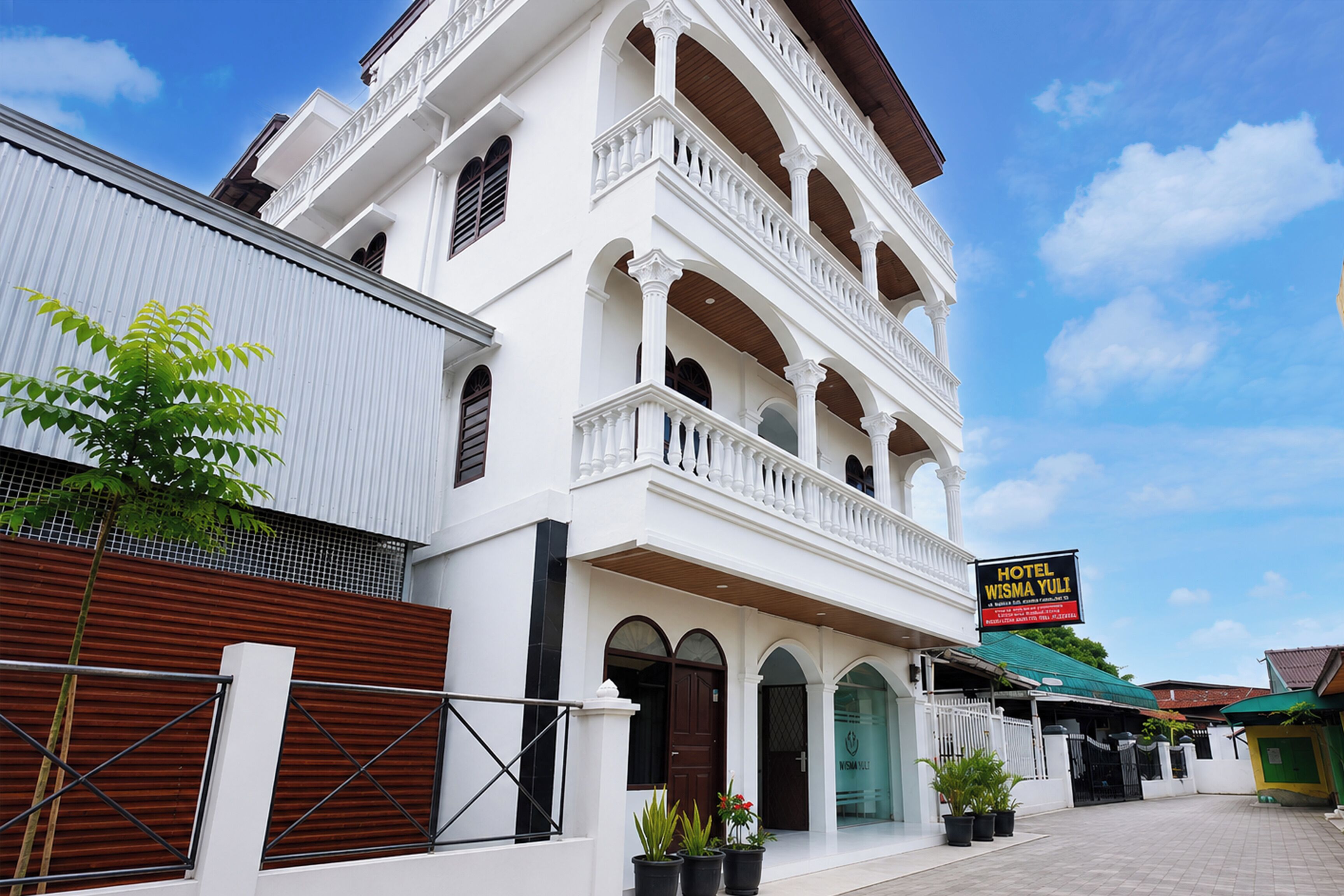 Um edifício branco de vários andares com varandas, uma placa com o nome Hotel Wisma Yuli, e vasos de plantas em frente.