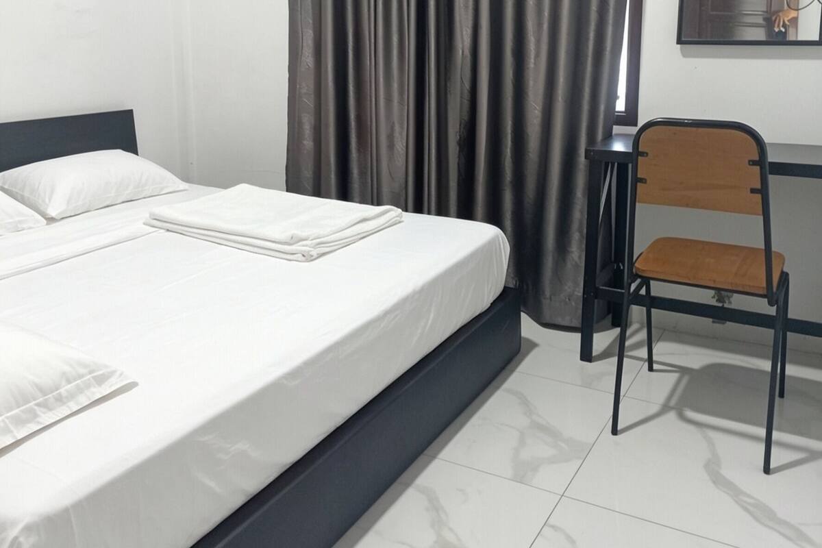 A neatly made bed with white linens, a black metal headboard, a wooden chair with a cushion, and a grey curtain.
