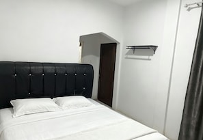 Een bed met een zwart hoofdeinde, wit beddengoed, een klein plankje en een deur op de achtergrond.