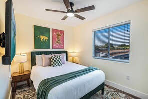 A hotel room with a bed, two wall-mounted lamps, a ceiling fan, a window with blinds, and two artwork pieces.