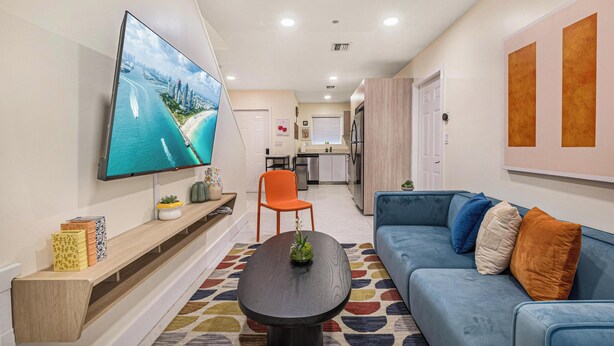 A modern living room with a flat-screen TV, a blue sofa, a coffee table, and a kitchen in the background. - 3BR Ft Lauderdale Townhome + Game Room + Parking (Fort Lauderdale)