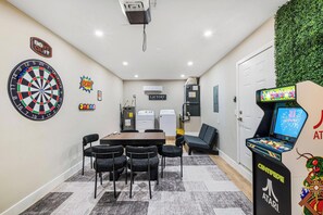 Ein Zimmer mit Dartscheibe, Tisch und Stühlen, einer Couch, einer Atari-Spielekonsole und einer Waschgelegenheit.
