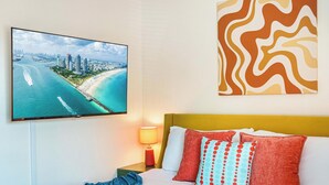 A hotel room with a bed, a TV displaying a coastal cityscape, a nightstand with a lamp, and a wall art piece. - 3BR Ft Lauderdale Townhome + Game Room + Parking (Fort Lauderdale)