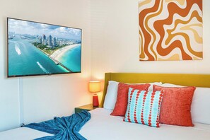 Quarto de hotel com uma cama, uma televisão exibindo uma paisagem costeira, uma mesinha de cabeceira com abajur e uma obra de arte na parede.