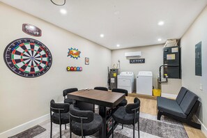 A room with a dartboard, a table, chairs, a couch, a laundry machine, and a sign that says 'LAUNDRY'.