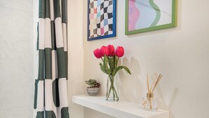 A bathroom with a white shelf holding a vase with red flowers, a small potted plant, and a reed diffuser. - 3BR Ft Lauderdale Townhome + Game Room + Parking (Fort Lauderdale)