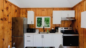 A kitchen with wooden paneling, white cabinets, a stainless steel refrigerator, a stove, and a window with a view of trees. - 2-Bedroom Cottage #4 (Nobel)