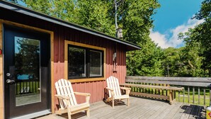 A wooden deck with two Adirondack chairs, a wooden bench, and a door with a window. - 2-Bedroom Cottage #4 (Nobel)