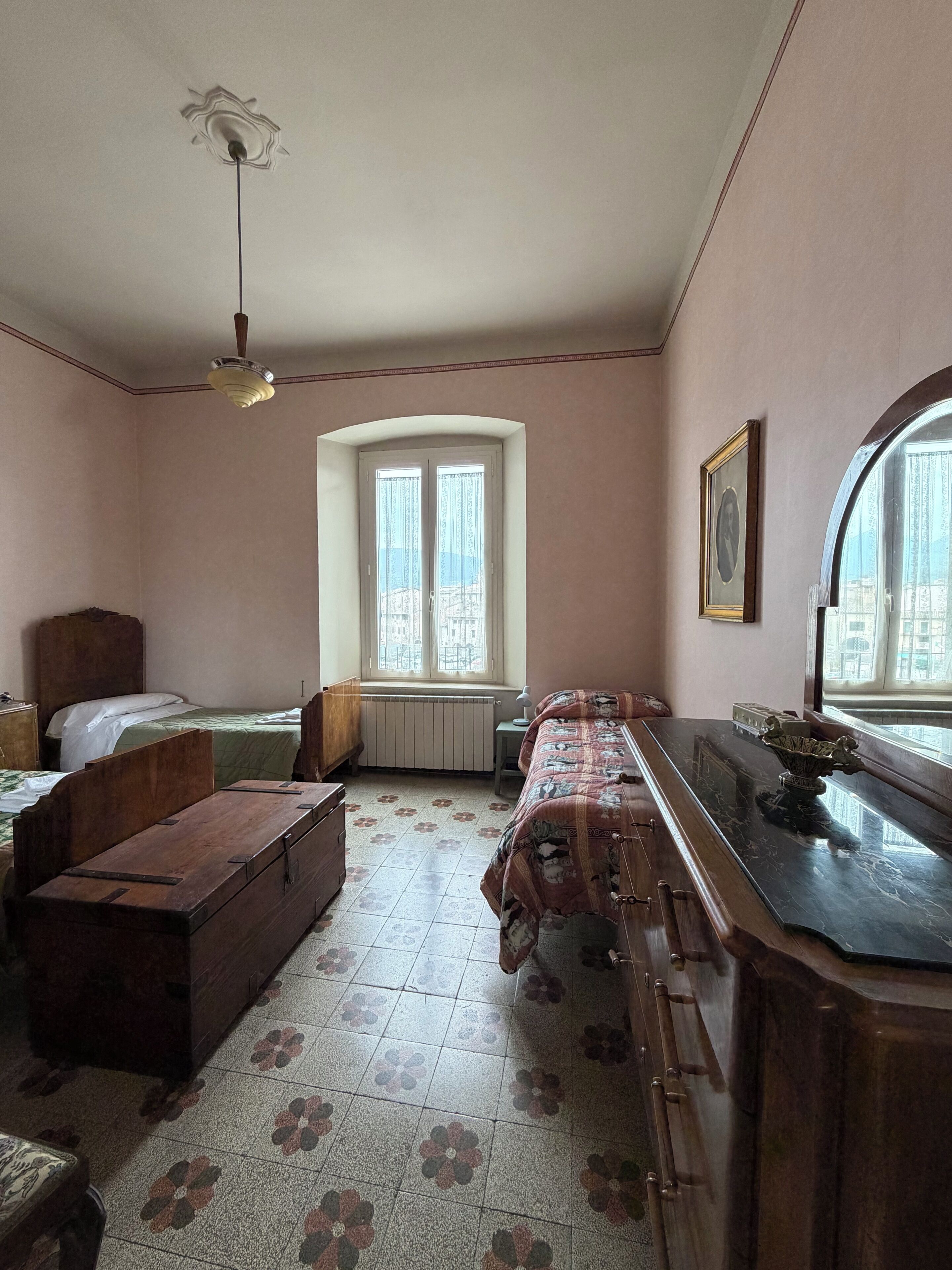 Quarto com cama, um baú de madeira, escrivaninha, radiador, janela com vista e ventoinha no teto.