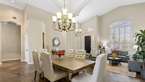 A spacious dining area with a wooden table, multiple chairs, a chandelier, and a large window with shutters.