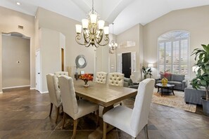 A spacious dining area with a wooden table, multiple chairs, a chandelier, and a large window with shutters. - Ahwatukee Foothills Home | Heated Pool | Grill (Phoenix)
