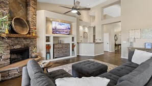 A spacious living room with a stone fireplace, a large flat-screen TV, a ceiling fan, and a modern kitchen in the background.