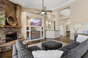 A spacious living room with a stone fireplace, a large flat-screen TV, a ceiling fan, and a modern kitchen in the background. - Ahwatukee Foothills Home | Heated Pool | Grill (Phoenix)