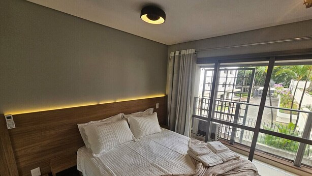Hotellihuone, jossa on suuri sänky, yöpöytä, sängynpääty, ikkuna verhoilla ja näkymä ulos.