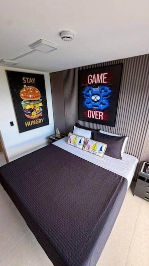A hotel room with a large bed, a flat-screen TV, a poster on the wall, and a small table with a lamp.