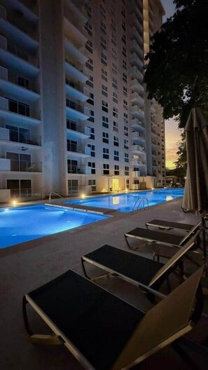 A swimming pool with lounge chairs, a tall building in the background, and a sunset sky.