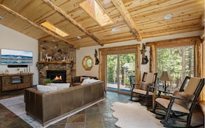 A log cabin interior with a stone fireplace, a large sofa, a leather ottoman, and a view of the outdoors through a sliding glass door. - Family Cabin, 2 Game Rooms, Basketball Court, Spa (Big Bear Lake,)