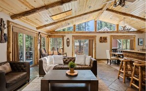 A rustic interior with a wooden ceiling, a leather sofa, a dining area, and large windows. - Family Cabin, 2 Game Rooms, Basketball Court, Spa (Big Bear Lake,)