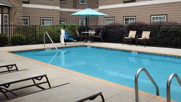 A swimming pool with lounge chairs and an umbrella, surrounded by a fence and a building with multiple windows.