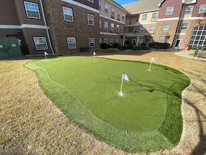 Un campo de golf artificial bien cuidado con banderas blancas que marcan los hoyos, rodeado por un edificio de ladrillo con numerosas ventanas.